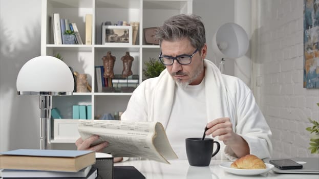 50s man having breakfast drinking morning coffee at home. Businessman checking newspaper for business and stock financial news.