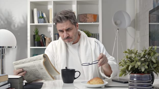 50s man having breakfast drinking morning coffee at home. Businessman checking newspaper for business and stock financial news.