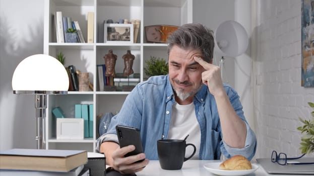 50s man having breakfast checking news on phone drinking morning coffee at home. Confident happy smiling.