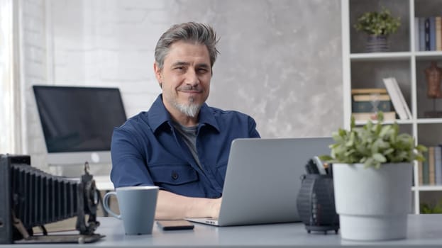 Bearded man working online with laptop computer at home sitting at desk. Businessman in home office, browsing internet. Portrait of mature age, middle age, mid adult man in 50s.