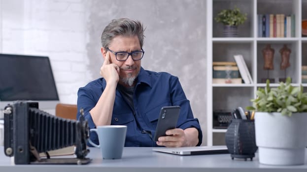 Bearded man working online with laptop computer and smart phone at home sitting at desk. Businessman in home office. Portrait of mature age, middle age, mid adult man in 50s.