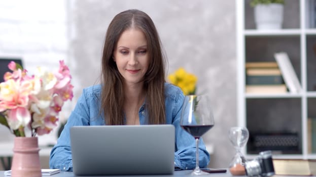 Young woman working online with laptop computer at home sitting at desk. Businesswoman in home office, browsing internet, online wine tasting.