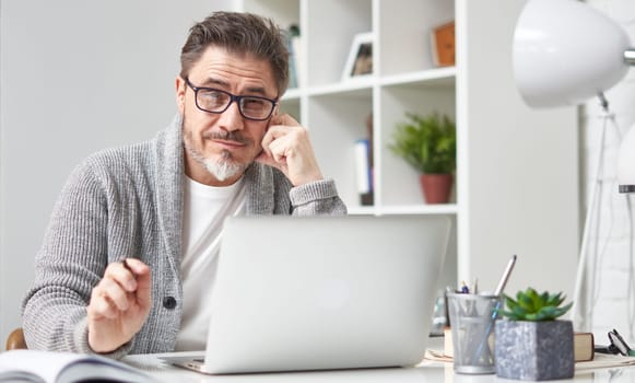 Older man working online with laptop computer at home sitting at desk. Home office, browsing internet, study room.
