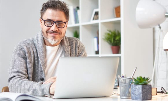 Happy older man working online with laptop computer at home sitting at desk. Home office, browsing internet, study room. Portrait of mature age, middle age, mid adult man in 50s.