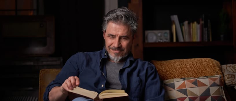 Happy man sitting on couch at home in cosy room reading book. Relaxing, leisure, education. ..Portrait of mature age, middle age, mid adult man, bearded, smiling, authentic look.