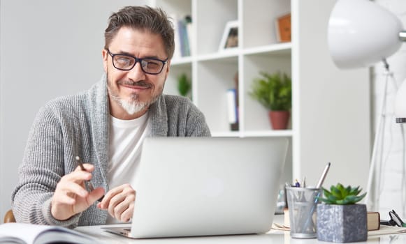 Happy older man working online with laptop computer at home sitting at desk. Home office, browsing internet, study room. Portrait of mature age, middle age, mid adult man in 50s.