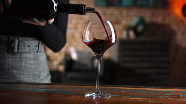 Elegant woman pouring red wine from wine bottle to wine glass at home in a cosy dark room.