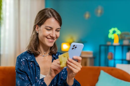 Happy young woman doing payments online purchase shopping in internet store. Smiling Caucasian girl holding banking credit card involved in verification process in mobile application on sofa at home.