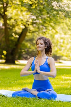 Young woman sitting on mat in lotus position, relaxing in park on summer day. Sporty carefree adult girl practicing yoga meditation, receives positive energy, healthy lifestyle. Active sportswoman.