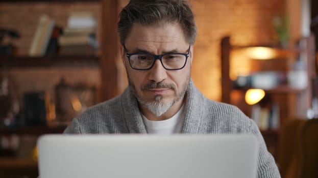 Older man working online with laptop computer at home sitting at desk. Home office, browsing internet, study room. Portrait of mature age, middle age, mid adult man in 50s.