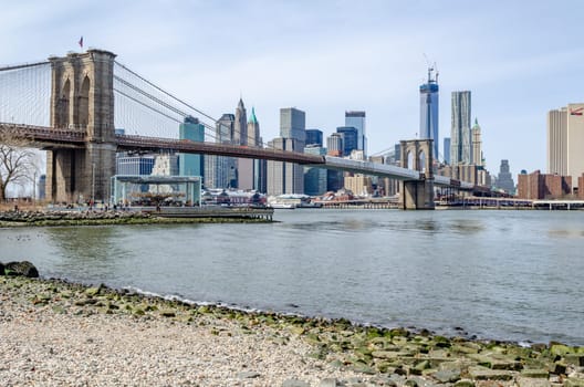 Brooklyn Bridge New York City with construction area on the bridge with Manhattan Skyline and One World Trade Center Construction area in the Background, Hudson river, beach and carousel in the forefront, wide angle shot, daytime with great weather, horizontal