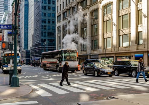 People crossing the Street on a crosswalk in Manhattan at East 48th Street, Cars and two busses waiting in the background, Smoke rises on the street behind a bus,  New York City during daytime, horizontal