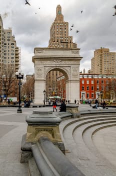 Washington Square Arch, New York City during a cold winter day with empty fountain in forefront, Birds flying over the building, people walking in the park, vertical