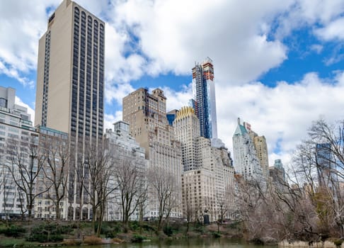 Skyline of Manhattan with Essex House and other Skyscraper, The Pond at Central Park Lake in forefront, view from low angle, cloudy sky during winter, horizontal