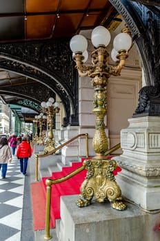 Entrance of The Plaza Hotel, New York City with golden Street Lamps and a red carpet in front of the Building, View from the side, People walking on the sidewalk in the background, vertical