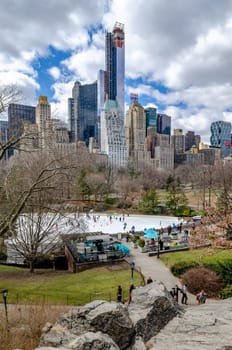 Wollman Rink with People Ice Skating during daytime in winter, view from the distance, Central Park New York City, benches and people walking in the forefront, rocks in the forefront, vertical