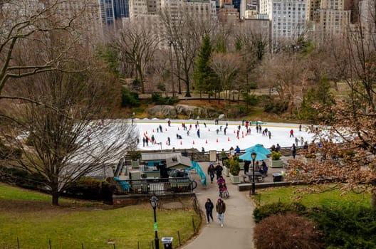 Wollman Rink with People Ice Skating during daytime in winter, view from the distance, Central Park New York City, benches and people walking in the forefront, horizontal