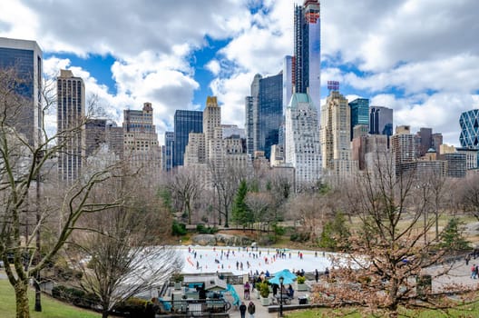 Trumps Wollman Rink with People Ice Skating during daytime in winter, view from the distance, Skyline of Manhattan in the Background, Central Park New York City, benches and people walking in the forefront, horizontal