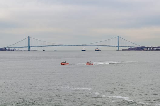 Two red United States Coast Guard ships and two blue and white industrial ships on Hudson river in front of Verrazano-Narrows Bridge, New York City, during winter evening with overcast, horizontal