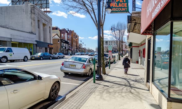 Retail Stores at Center of the City near Cliffside Park, New Jersey, old woman walking on the sidewalk, cars parked next to the street, during a sunny winter day, horizontal
