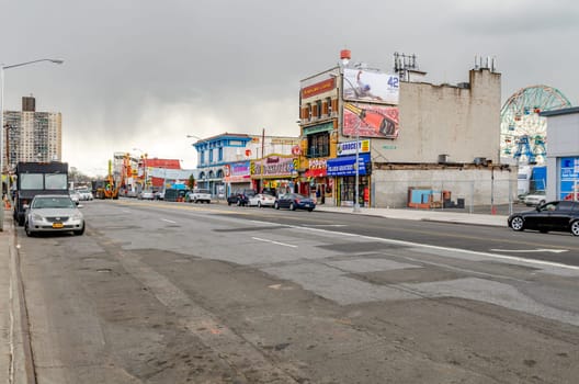 City Street with retail stores in Coney Island, ferris wheel behind the stores, cars parked next to the street, New York City during winter with overcast, horizontal