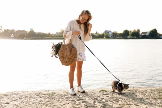 Young blonde, woman, girl walking on the beach in spring or autumn in a dress and white sweater with a Scottish straight kitten on a sunny day, sunset