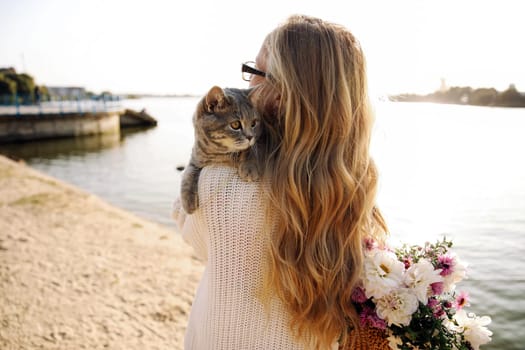 Young blonde girl with glasses holds a cat breed Scottish Straight. Walk with pet near the lake, river at sunset on the beach