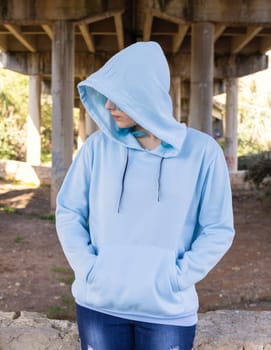 Teenager wearing light blue oversize hoodie and jeans looking aside and keeping hands in pocket. Blue haired teen girl stays outdoors against bridge pillars. Clothing mockup