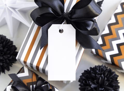 Present with vertical blank gift tag and black bow close up. Gift boxes wrapped in Striped geometric paper near black and white decor. Christmas, New Year, Birthday, Anniversary label mockup