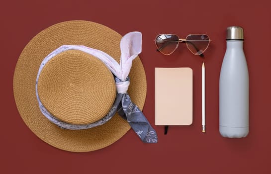 Straw hat, notebook, pensil, heart sunglasses and reusable bottle top view on red. Female summer clothes and accessories flat lay. Hardcover textbook and bottle mockup. Travel concept