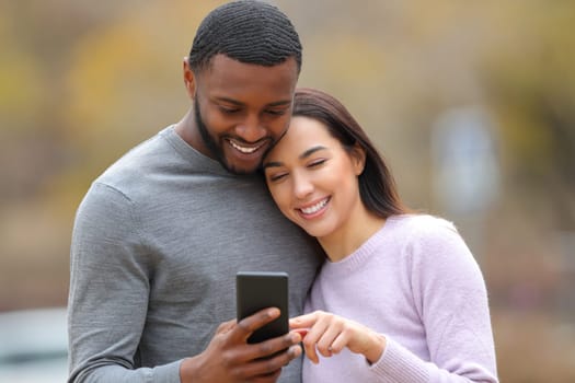 Happy interracial couple checking smart phone outdoors