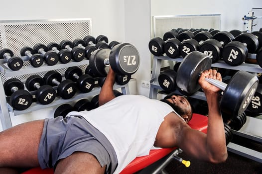 An African-American man does a dumbbell press while lying on a bench in the gym. High quality photo