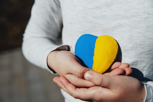 Small little kid hold heart shaped stone with Ukraine blue-yellow national flag or anthem. Girl love native country. Child stand for Ukrainians. Stop war, agression, invation. People agains the war.