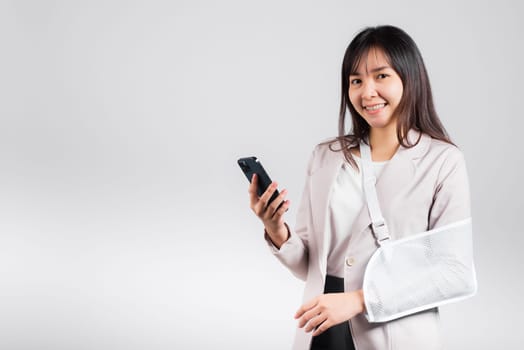 Woman confident smiling broken arm after accident and wear arm splint for treatment and hold smartphone, happy Asian female sling supported hand isolated on white background, social media, copy space