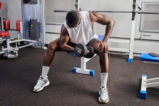 An African American man shakes his biceps while sitting on a bench in the gym. High quality photo
