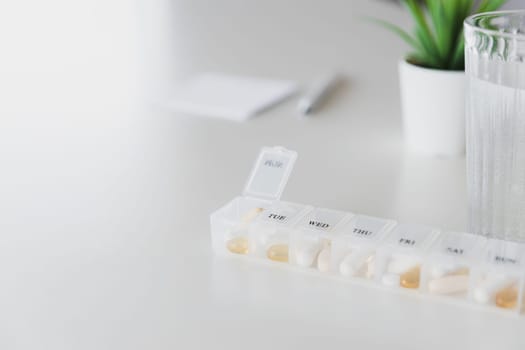 Closeup of medical pill box with doses of tablets for daily take a medicine with white, yellow drugs and capsules used for treatment, cure the disease. Glass cup of water on table, gray background.