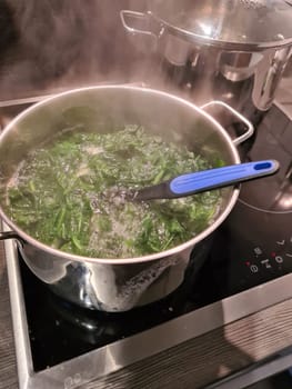 Spinach cooking preparation. Stewed spinach in black cooking pot with spoon on rustic background with ingredients and kitchen utensils . Top view. Home cuisine. Healthy food nutrition