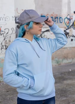 Blue haired Teenage girl in blue hoodie and baseball cap touching her cap and looking right. Blue haired teen girl stays outdoors against graffiti wall. Clothing and cap mockup