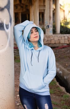 Smiling Teenager wearing light blue oversize hoodie and jeans stays under bridge . Blue haired teen girl stays outdoors against bridge pillars. Clothing mockup