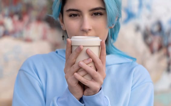 Teenage girl in light blue oversize hoodie with coffee to go and looking stright.Blue haired teen girl staying outdoors near graffiti wall, head shot. Hipster and adolescence concept