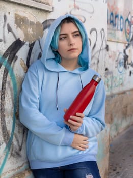 Teenager in blue hoodie with red water bottle looking aside. Blue haired teen girl staying outdoors near graffiti wall. Clothing and reusable bottle mockup. Hipster and adolescence concept