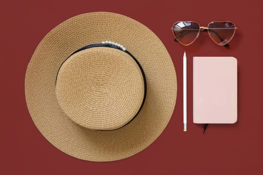 Straw hat, notebook, heart sunglasses and pensil top view on red background. Hardcover textbook mockup. Female summer clothes and accessories flat lay. Travel concept