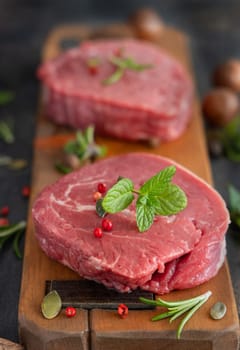 Raw beef fillet steaks on wooden board on dark background close up. Portioned filet mignons with spices and herbs. Concept of summer food