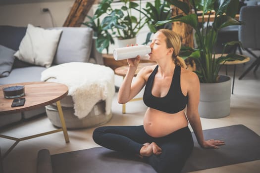 Young happy and cheerful beautiful pregnant woman taking a break, hydrating, drinking water from the botle after home well being workout program