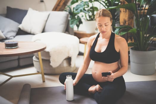 Young happy and cheerful beautiful pregnant woman taking a break, hydrating, drinking water from the botle after home well being workout program