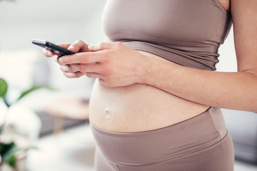 Closeup photo of pregnant female belly. Woman holding and using mobile smart phone app at home interiors. Pregnancy, technology, online shopping, preparation and expectation concept.