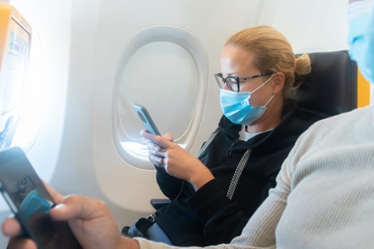 A young woman wearing face mask, using smart phone while traveling on airplane. New normal travel after covid-19 pandemic concept