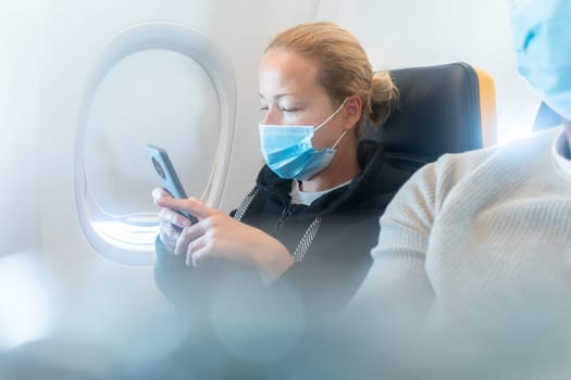 A young woman wearing face mask, using smart phone while traveling on airplane. New normal travel after covid-19 pandemic concept