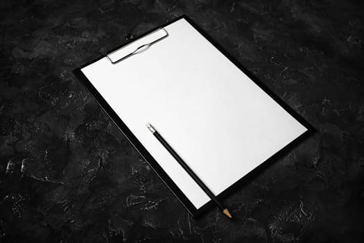 Clipboard with blank paper and pencil on black plaster background.