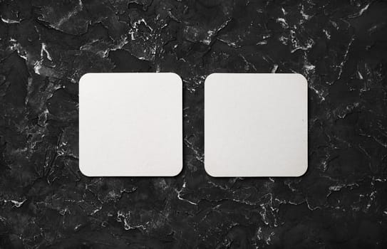 Photo of two blank square beer coasters on black plaster background. Blank template for your design. Top view. Flat lay.